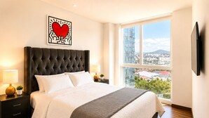 Ein modernes Hotelzimmer mit einem großen Bett, Blick auf die Stadt, einem Flachbildfernseher und einem Wandbild mit Herzmotiv.
