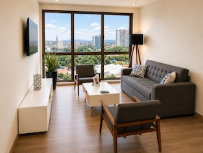 Un salon moderne comprenant un canapé, des fauteuils, une table basse, un téléviseur et une grande fenêtre donnant sur la ville.