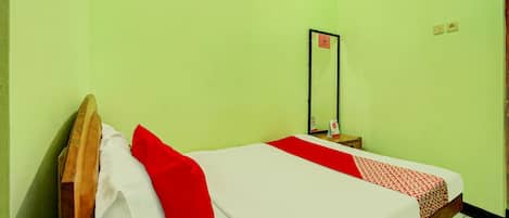 Una cama individual con cabecera de madera, ropa de cama blanca y roja, y una manta roja. Se ve una mesita de noche pequeña con un espejo y un objeto rojo.