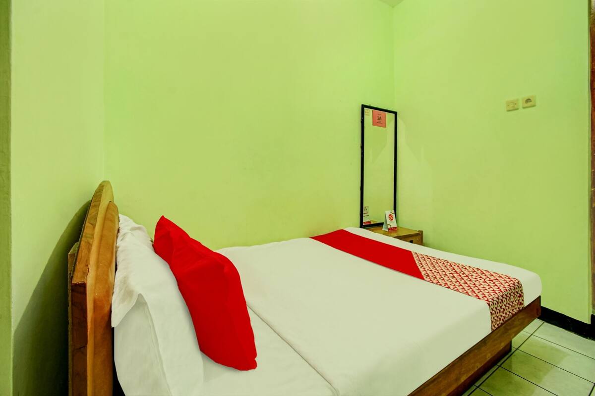 Uma cama de solteiro com cabeceira de madeira, roupa de cama branca e vermelha, e um cobertor vermelho. É visível uma pequena mesa de cabeceira com um espelho e um objeto vermelho.
