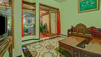 A room with a wooden sofa, a television, a patterned floor, and a view of a garden through a glass door.