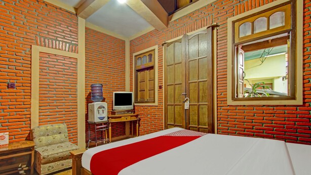 A hotel room with a brick-patterned wall, a bed with a red bedspread, a wooden wardrobe, a television on a wooden stand, and a small table with a chair. - OYO 90031 Villa Papada (Batu)