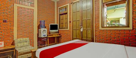 A hotel room with a brick-patterned wall, a bed with a red bedspread, a wooden wardrobe, a television on a wooden stand, and a small table with a chair.