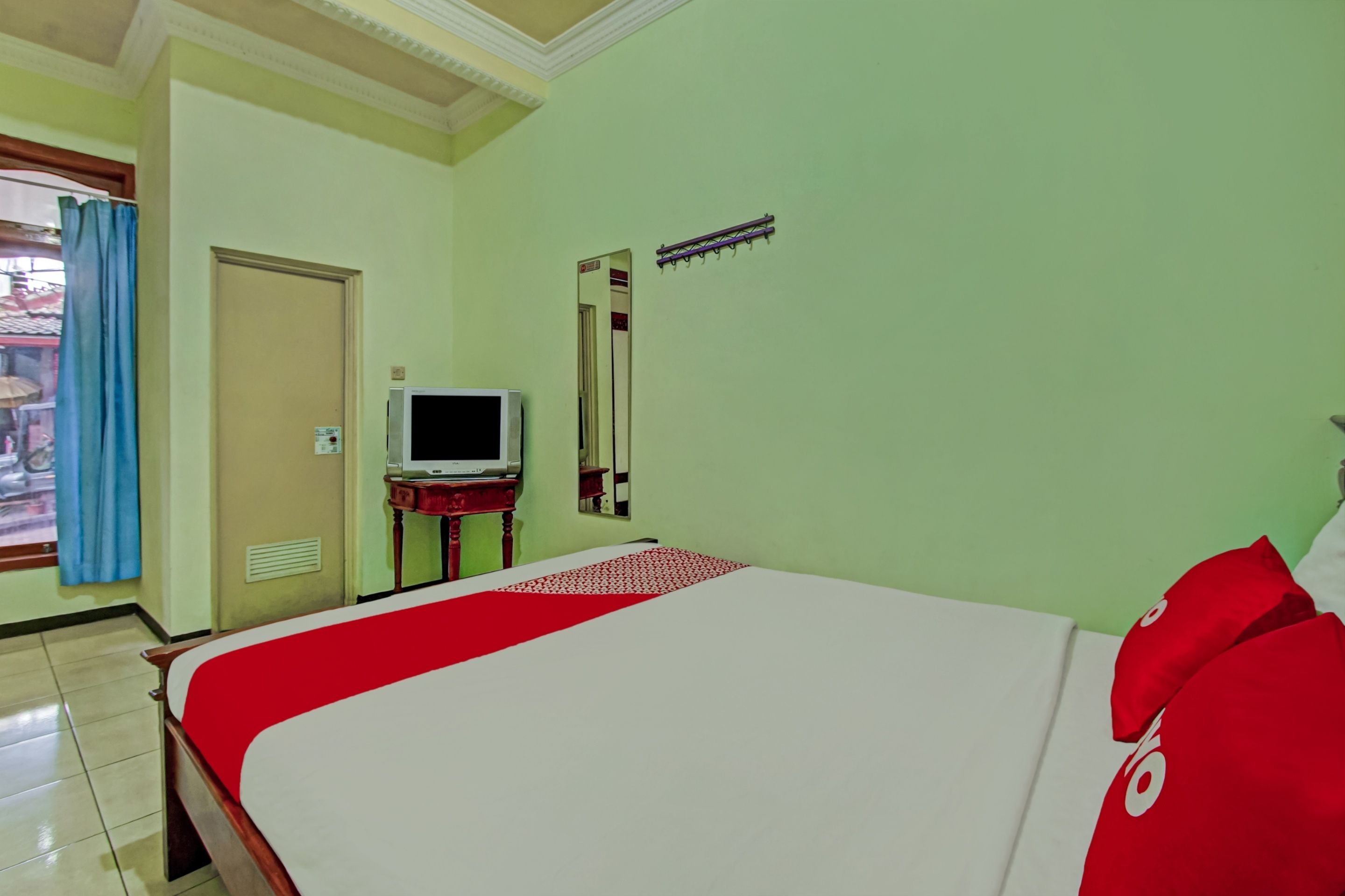 A hotel room with a bed, a television on a stand, a mirror, and a window with curtains.