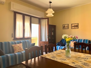 Una sala de estar con un sofá, una mesa de comedor con mantel floral y un florero con flores.