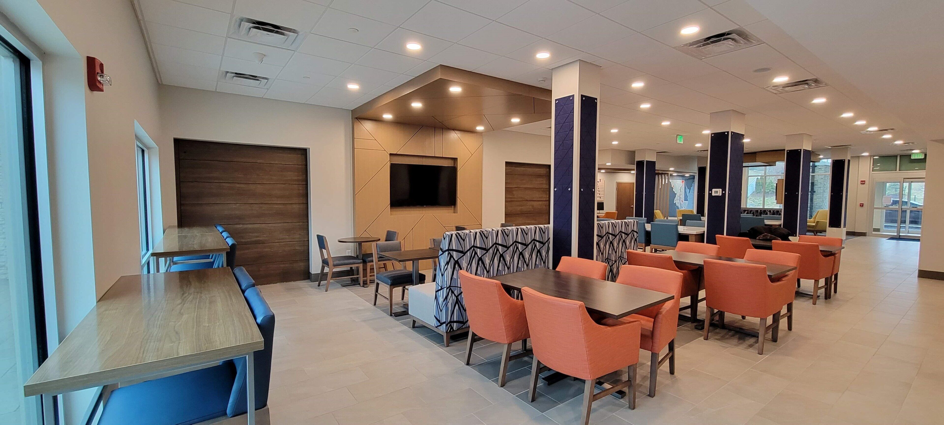 A modern restaurant with high tables and orange chairs, a large TV screen, and a wooden accent wall.