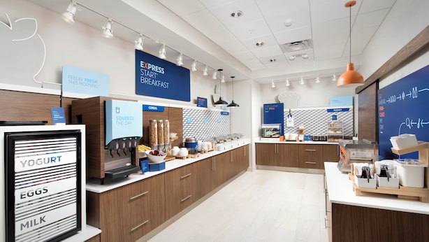 A modern breakfast area with labeled dispensers for yogurt, eggs, and milk, a coffee machine, and a chalkboard menu. - Holiday INN EXP White Marsh (WHITE MARSH)