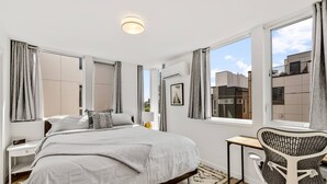 Ein modernes Schlafzimmer mit einem großen Bett, einem Schreibtisch und einem Stuhl. Vor den Fenstern hängen Vorhänge, und eine Klimaanlage ist an der Wand angebracht.
