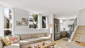 Ein modernes Wohnzimmer mit einem beigefarbenen Sofa, einem Holztisch und einer Küche im Hintergrund.