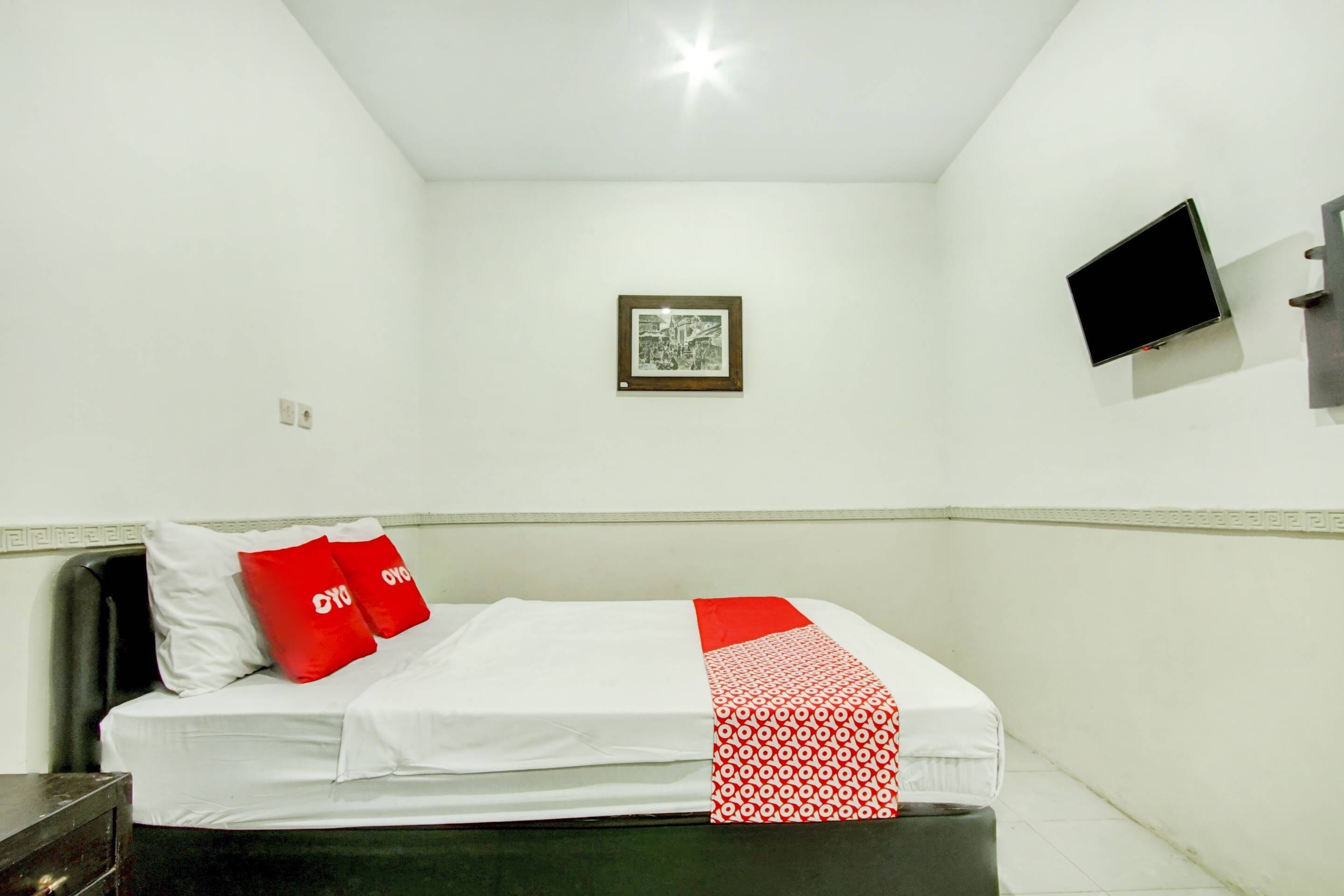 A single bed with a white and red bedspread, two red pillows, and a small framed picture on the wall.