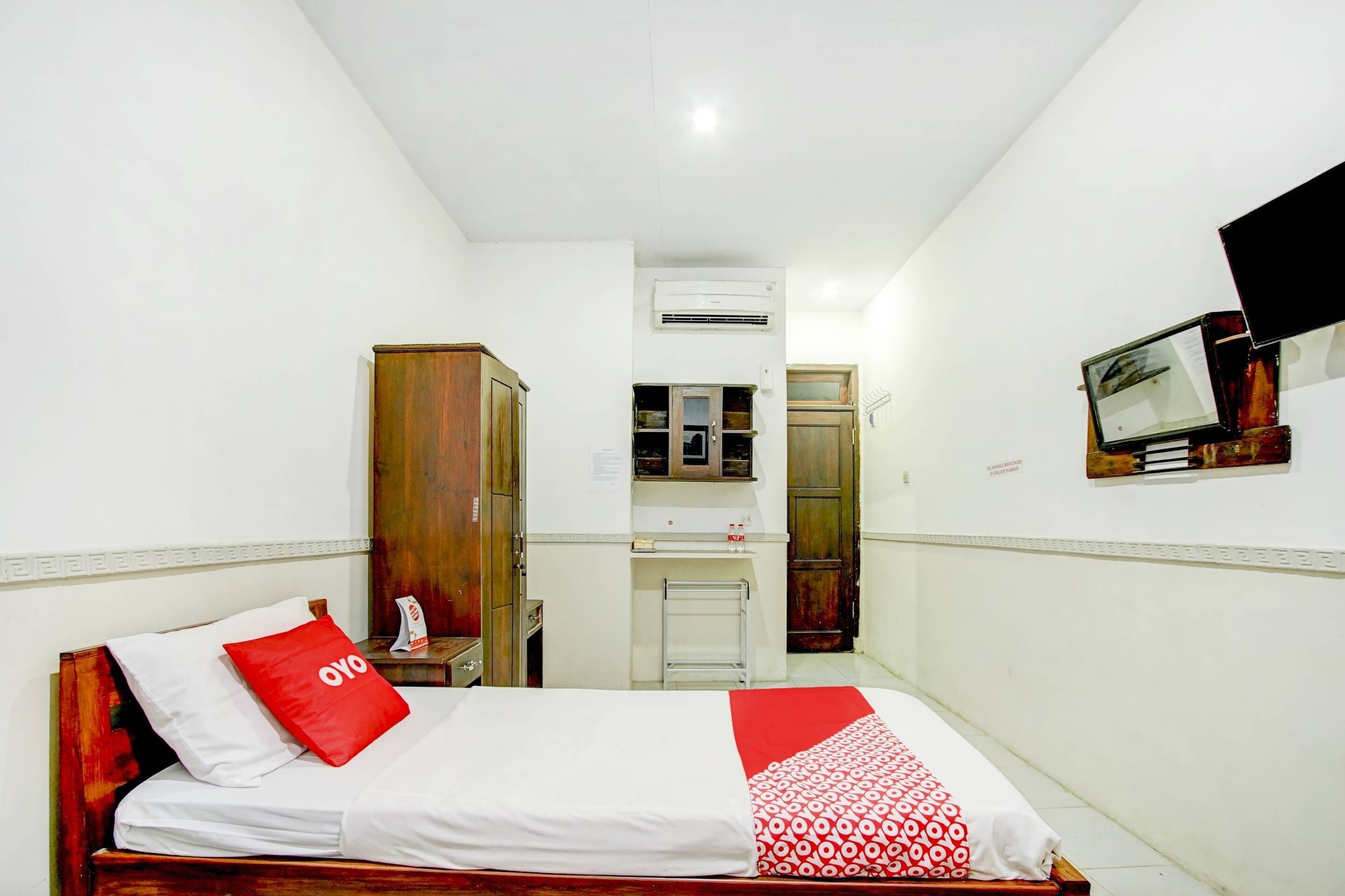 A small, clean room with a bed, a wooden wardrobe, a small desk with a chair, a mirror, and a television mounted on the wall.