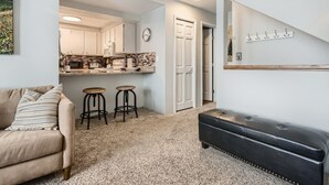 A modern living room with a sofa, a coffee table, and a black ottoman. There is a kitchen area with a countertop and stools visible through an open door. - Creekside Home | 3 BR/3BA | PVT Hot Tub (Frisco)