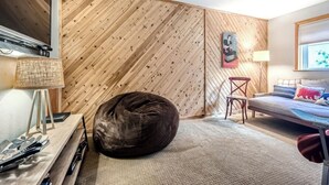 A modern room with a wooden slatted wall, a bed, a chair, a bean bag, and a desk with a lamp. - Creekside Home | 3 BR/3BA | PVT Hot Tub (Frisco)