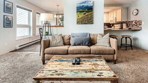 A living room with a beige sofa, a coffee table, and a painting of a mountain landscape. - Creekside Home | 3 BR/3BA | PVT Hot Tub (Frisco)