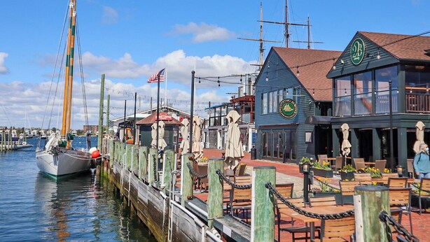 Um cais à beira-mar com um veleiro, um restaurante com mesas ao ar livre e um edifício com uma placa verde.