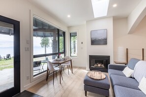 A modern living room with a fireplace, a dining area with a table and chairs, and large windows offering a view of the outdoors.