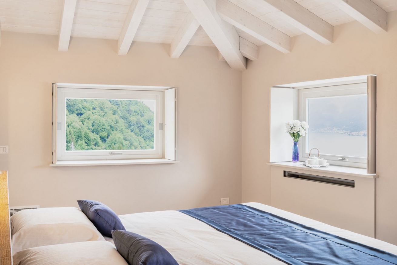 A bedroom with a bed, two windows, and a view of greenery and a cityscape.