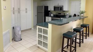 Cocina con gabinetes blancos, refrigerador de acero inoxidable, microondas y estufa. Hay tres taburetes de bar y un bote de basura.