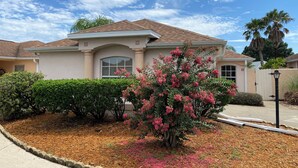 Una casa de un piso con fachada color crema, techo marrón y un árbol de flores rosadas en el jardín delantero.
