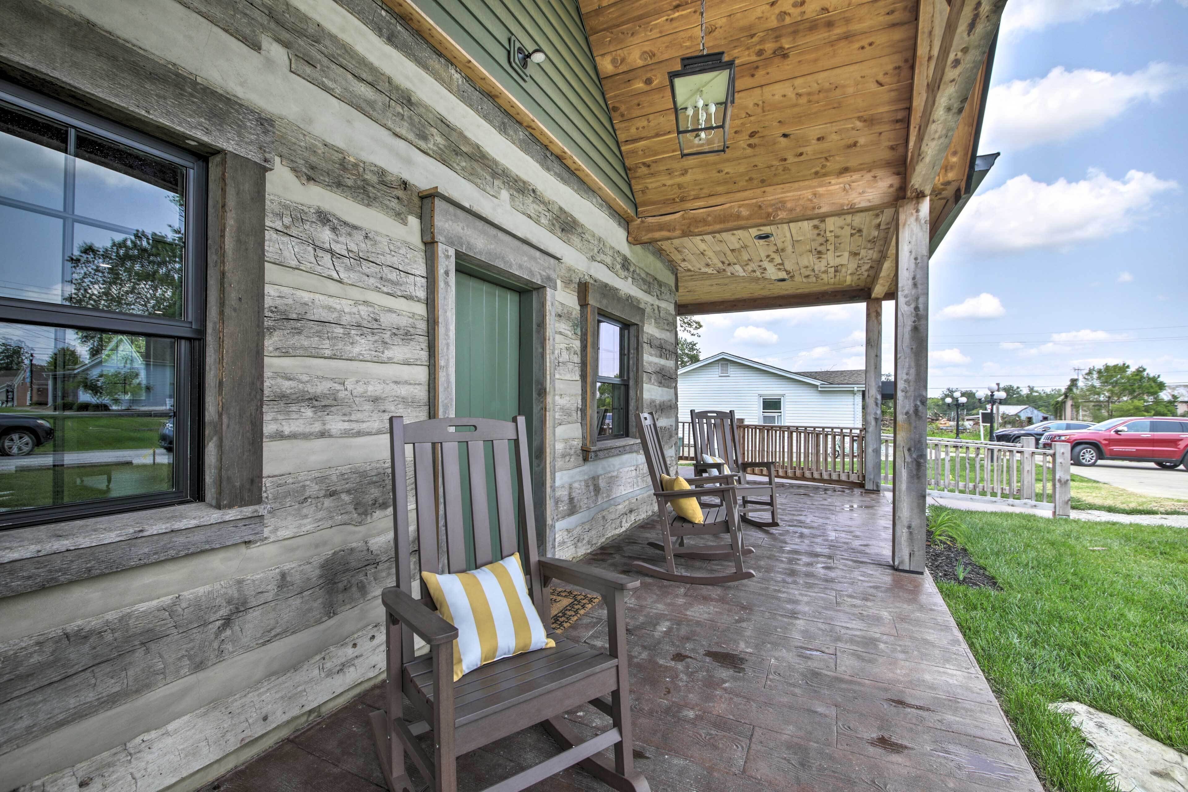 A wooden porch with rocking chairs and a bench, a green door, and a view of a white house in the background.