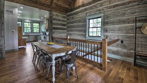 A rustic log cabin interior with a dining area featuring a wooden table and chairs, a kitchen with modern appliances, and a large window providing natural light. - ‘Bross Brother's Cabin’ w/ Wraparound Porch! (Cottleville)