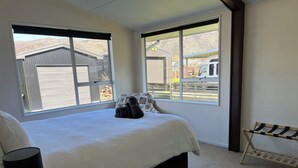 Un dormitorio con una cama grande, una mesita de noche con lámpara, un banco de madera y una ventana con vistas a un edificio y un vehículo estacionado.