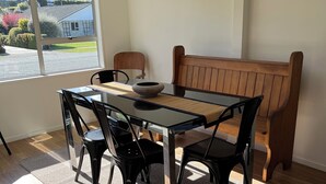 Un comedor con mesa de vidrio, sillas negras, un banco de madera y vista a una calle residencial a través de amplios ventanales.