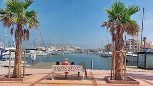 Marina con barcos, palmeras y un banco donde hay gente sentada.