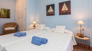 Une chambre d’hôtel avec deux lits, un fauteuil en osier et des œuvres d’art accrochées aux murs.