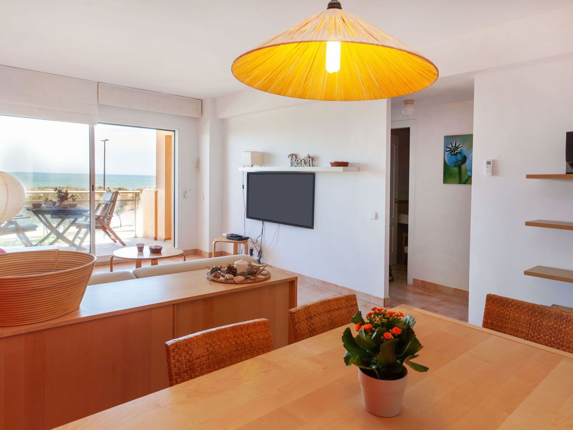 Un salon moderne doté d’une grande fenêtre, d’un coin repas avec une table et des chaises en bois, d’une lampe suspendue jaune et d’un téléviseur à écran plat fixé au mur.