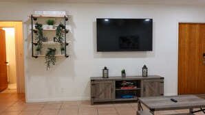 Ein Wohnzimmer mit an der Wand montiertem Fernseher, einem Holztischchen mit Topfpflanzen und einem gefliesten Boden.