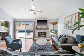 Uma sala de estar moderna com lareira, ventilador de teto e uma grande janela com vista para uma piscina.