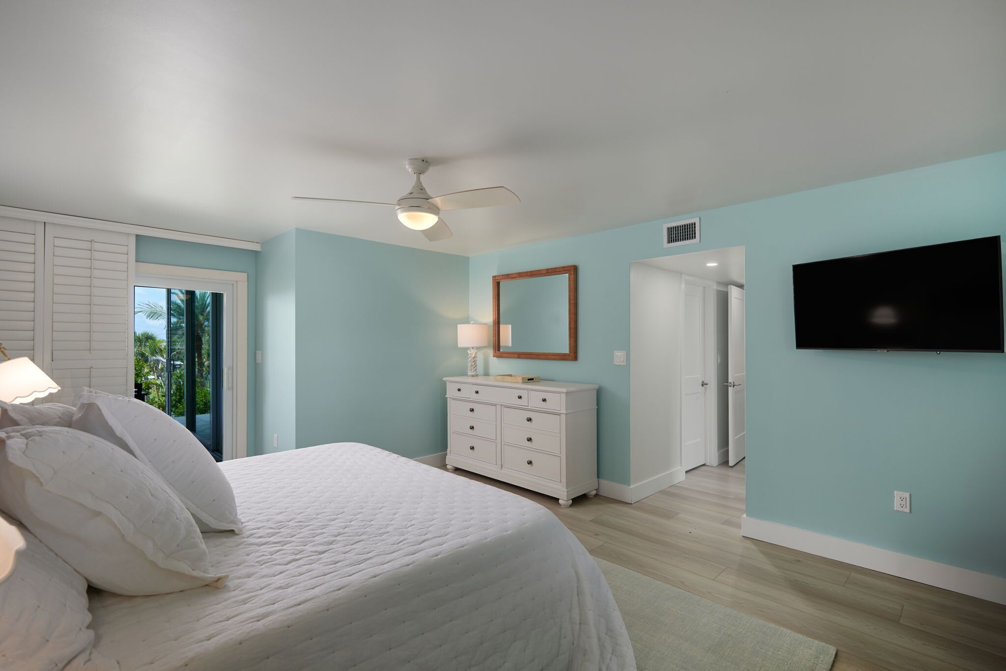 Une chambre à coucher comprenant un lit, une commode, un ventilateur de plafond, une télévision et une porte donnant sur une autre pièce.