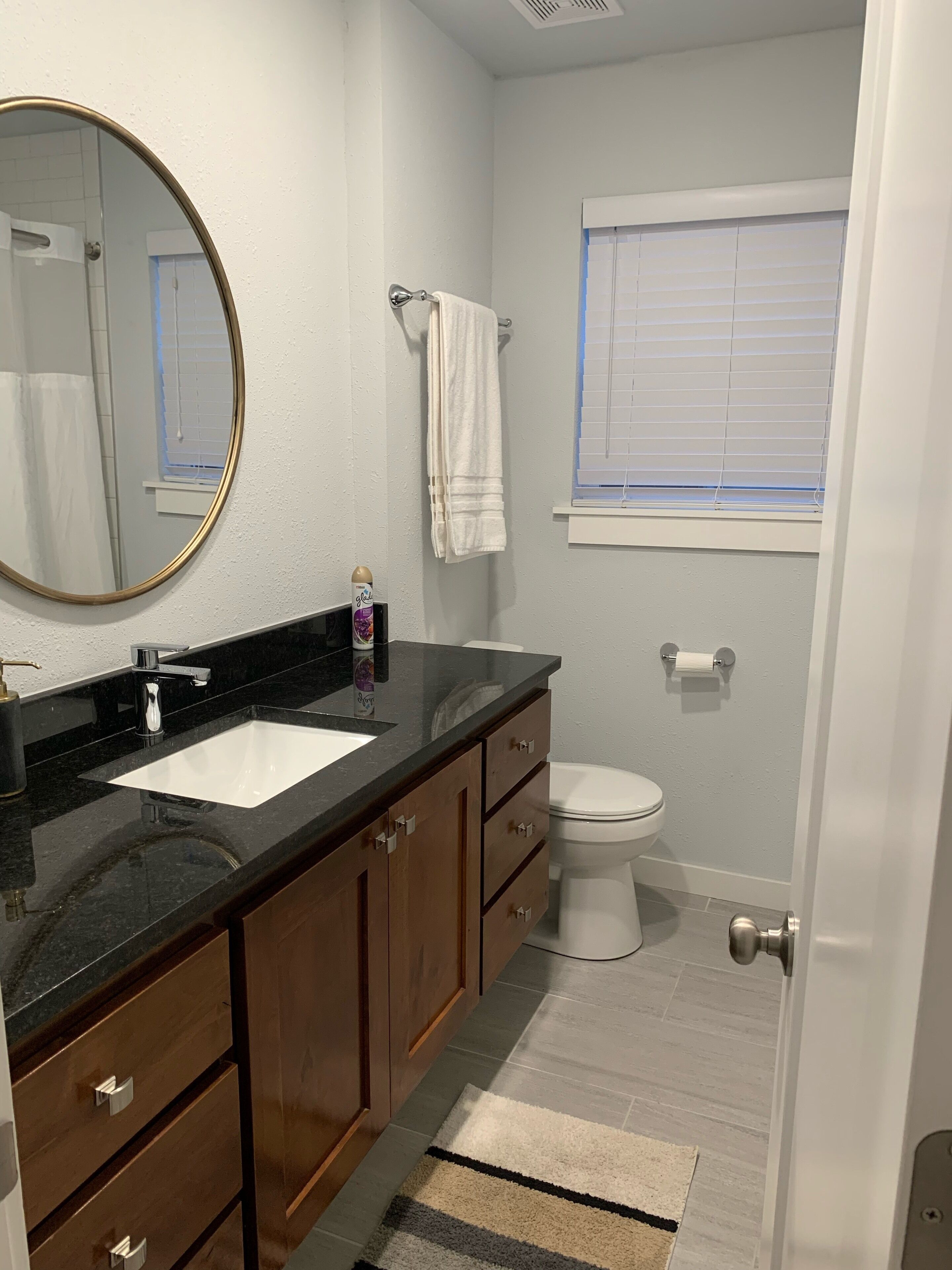 A bathroom with a black countertop sink, wooden cabinets, a white toilet, and a window with blinds.