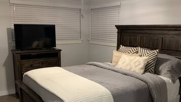 A bedroom with a bed, a television on a stand, a ceiling fan, and a window with blinds.