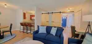 Una sala de estar moderna con un sofá azul, un comedor con una mesa redonda y sillas, y una cocina al fondo.