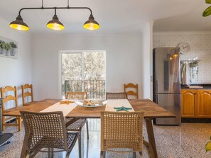A dining area with a wooden table and chairs, a kitchen with a refrigerator, and a window with a view of trees.