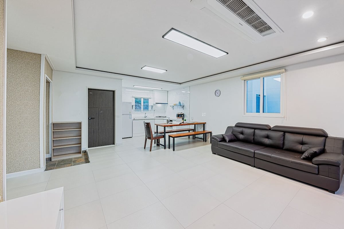A modern living room with a black leather sofa, a dining area with a wooden table and chairs, and a kitchen in the background.