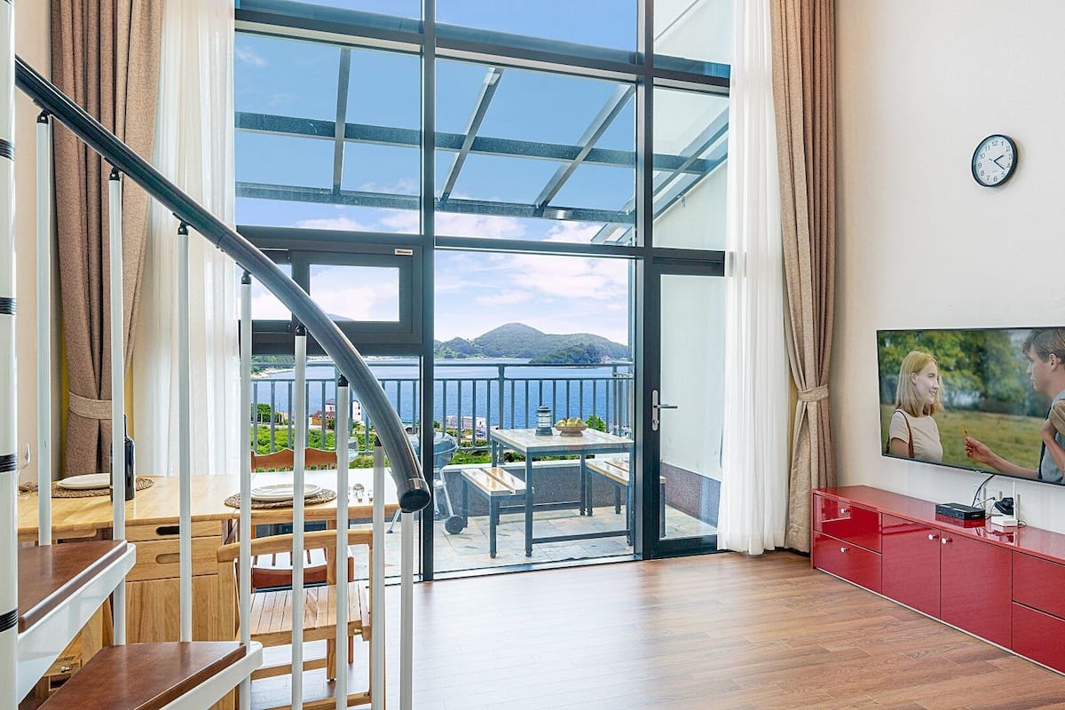 A modern living room with a wooden floor, a staircase, a balcony with a view of the sea, a red cabinet, and a wall-mounted TV.