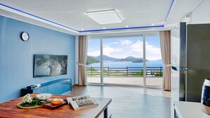 A modern room with a large window, a flat-screen TV, a wooden dining table, and a refrigerator. - Clover spa pension (Geoje)