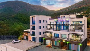 A multi-story hotel with balconies, a car parked in front, and a mountainous backdrop. - Clover spa pension (Geoje)