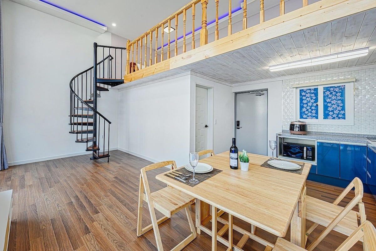 A modern interior with a wooden dining table, chairs, a spiral staircase, and a kitchen area with blue cabinets.