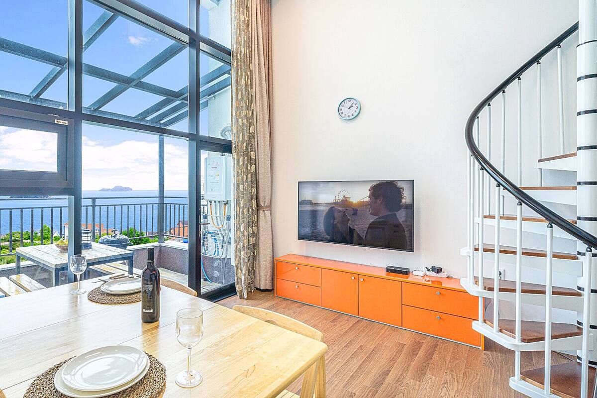 A dining area with a table set for two, a TV mounted on the wall, and a view of the sea through large windows.