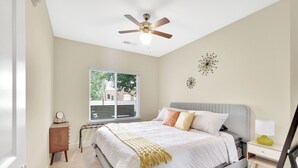Une chambre à coucher comprenant un lit, une table de chevet, une commode, un ventilateur de plafond, une fenêtre et des éléments de décoration murale.