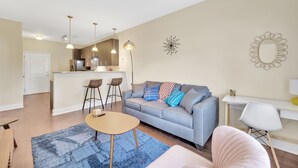 Un salon moderne avec un canapé gris, une table basse ronde et un tapis bleu. On y trouve également un coin cuisine équipé d’un réfrigérateur, d’un évier et d’une table à manger avec des chaises.