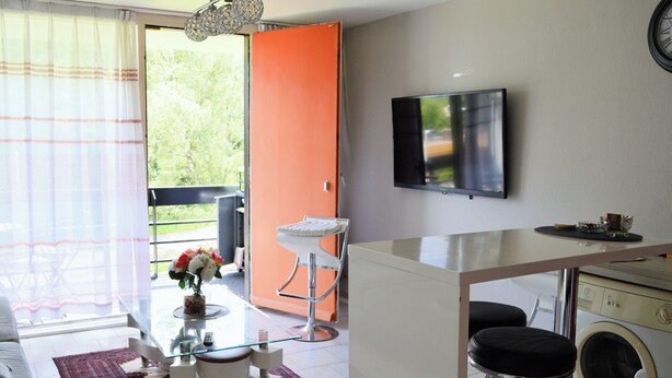 Un espace de vie compact comprenant un canapé, une table basse en verre, un petit coin cuisine avec un lave-linge et un téléviseur fixé au mur.