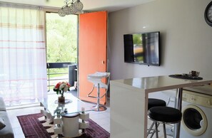 A compact living space with a sofa, a glass coffee table, a small kitchen area with a washing machine, and a TV mounted on the wall. - Peace and Green 19 (La Calamine)