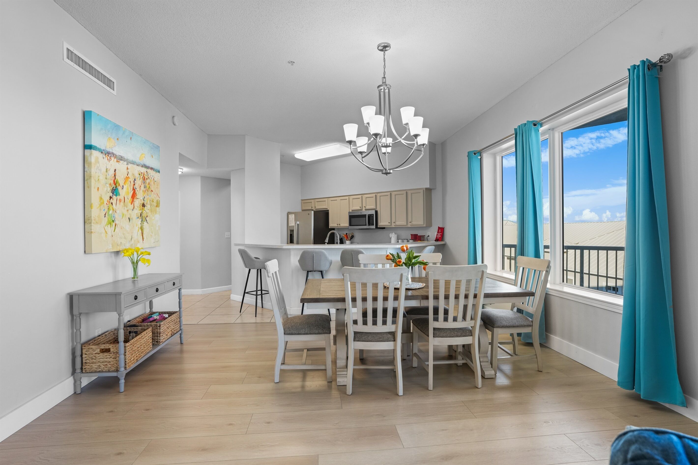 A modern kitchen with a central island, a dining area with a wooden table and chairs, and a view of the sky through large windows with teal curtains.