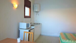 A small kitchen with a microwave, stove, and sink. There is a wooden table with a mug on it. A bed with a colorful blanket is visible in the corner. - Basic studio in Canyelles with communal terrace + private outdoor parking. (Roses)