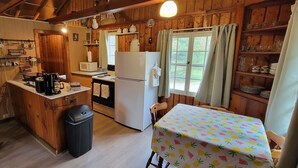 A cabin interior with a wooden interior, a kitchen area including a refrigerator, a microwave, and a coffee maker. There is a table with a fruit-patterned tablecloth and a chair. - Summer, Mermaid Coast Cottages on Lake Huron (East Tawas)